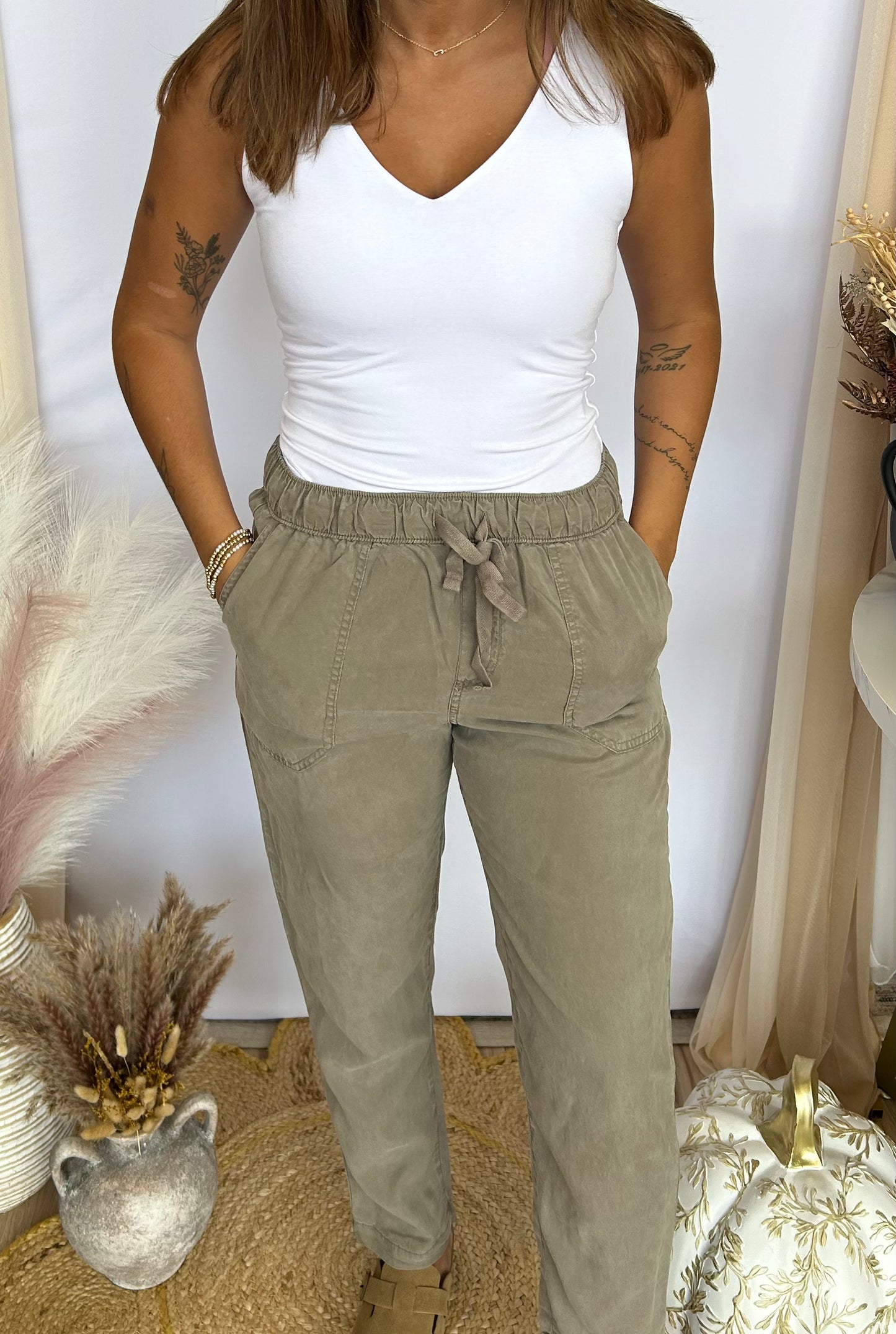 Person wearing a white tank top and beige pants in a decorative setting with plants and decor items.