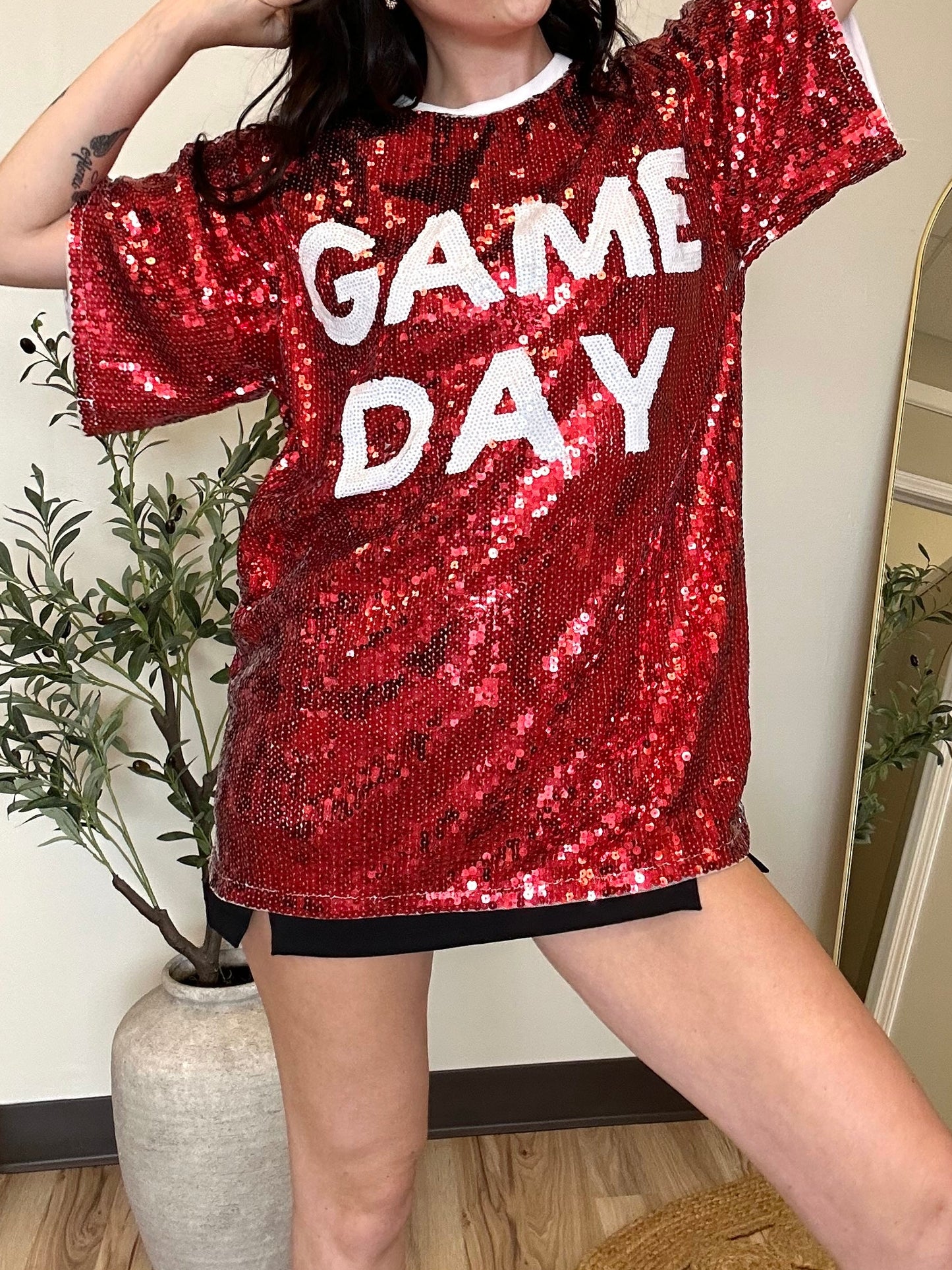 A person wearing a red sequin top with the text 'GAME DAY' written in large letters, posing in a room with a mirror and potted plants in the background.
