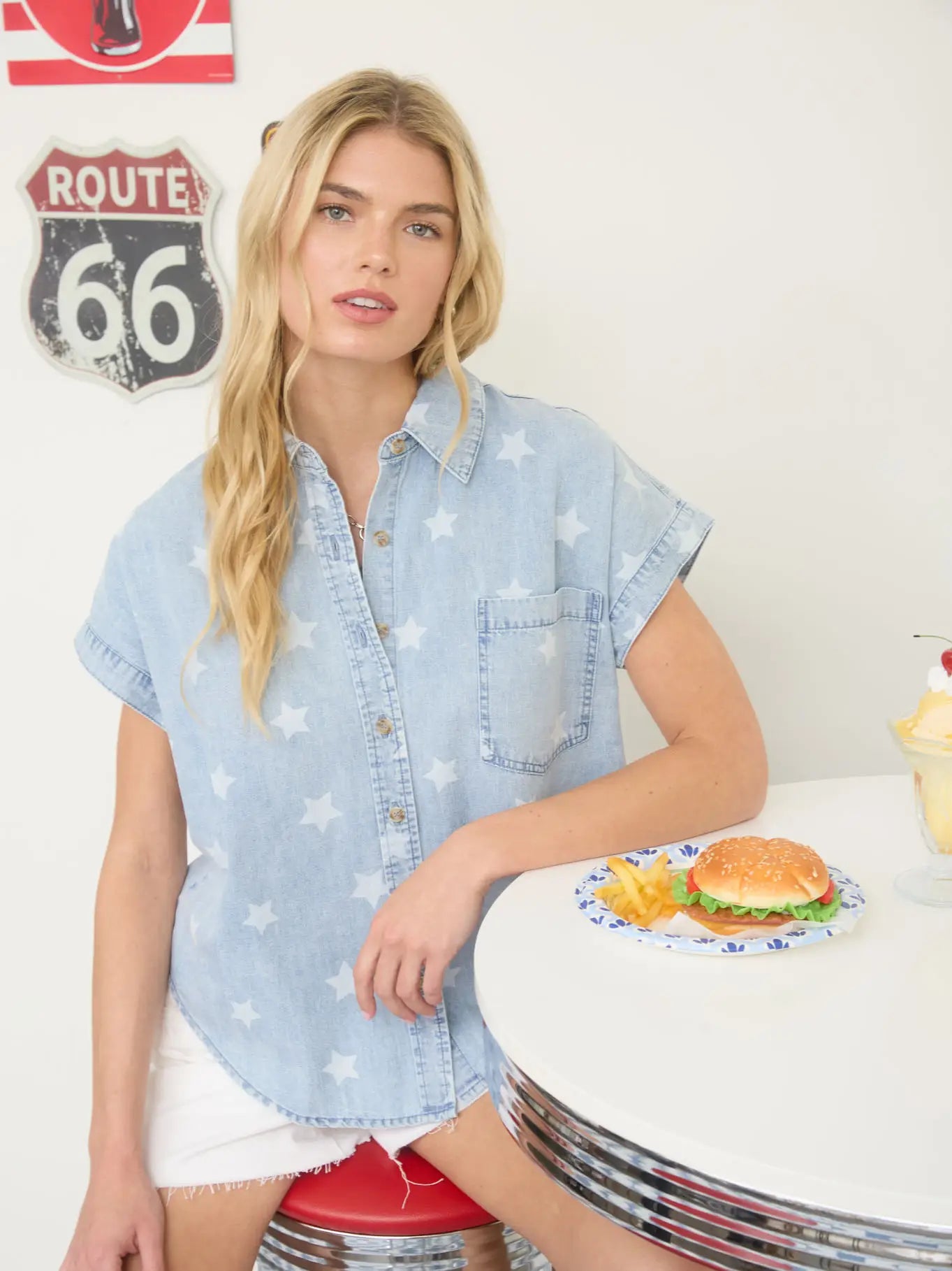 Woman in a denim shirt standing next to a table with a hamburger and fries, Route 66 sign in the background.
