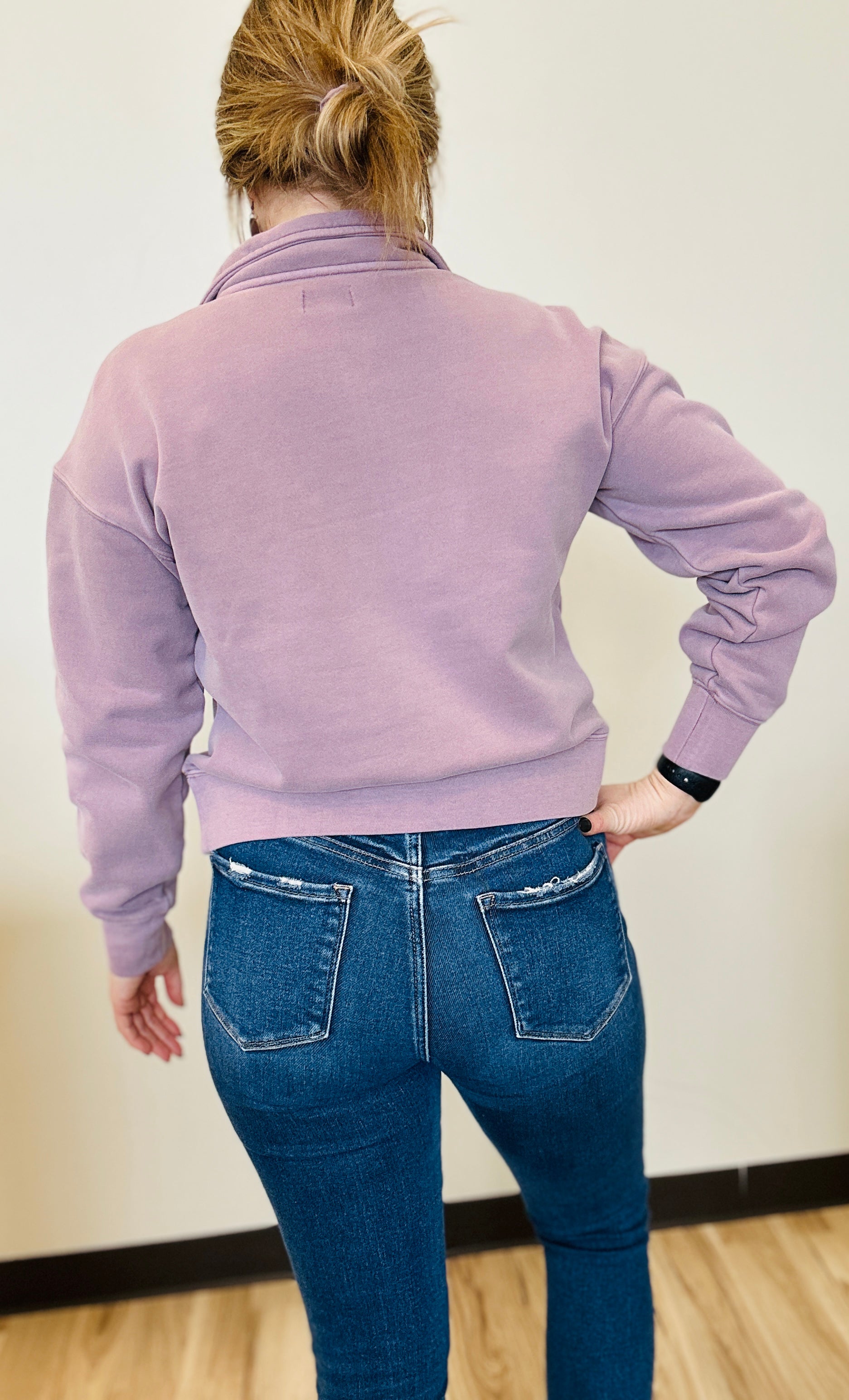Person wearing a light purple jacket and blue jeans standing against a plain wall.