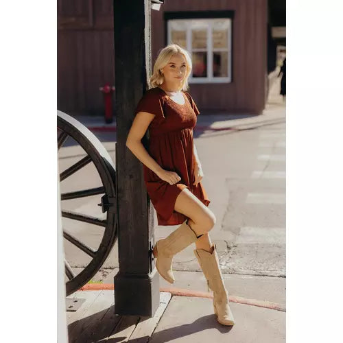 A woman wearing a short-sleeve rust-brown dress and beige cowboy boots leans against a dark wooden post on a sunlit street, with a building and wagon wheel in the background.
