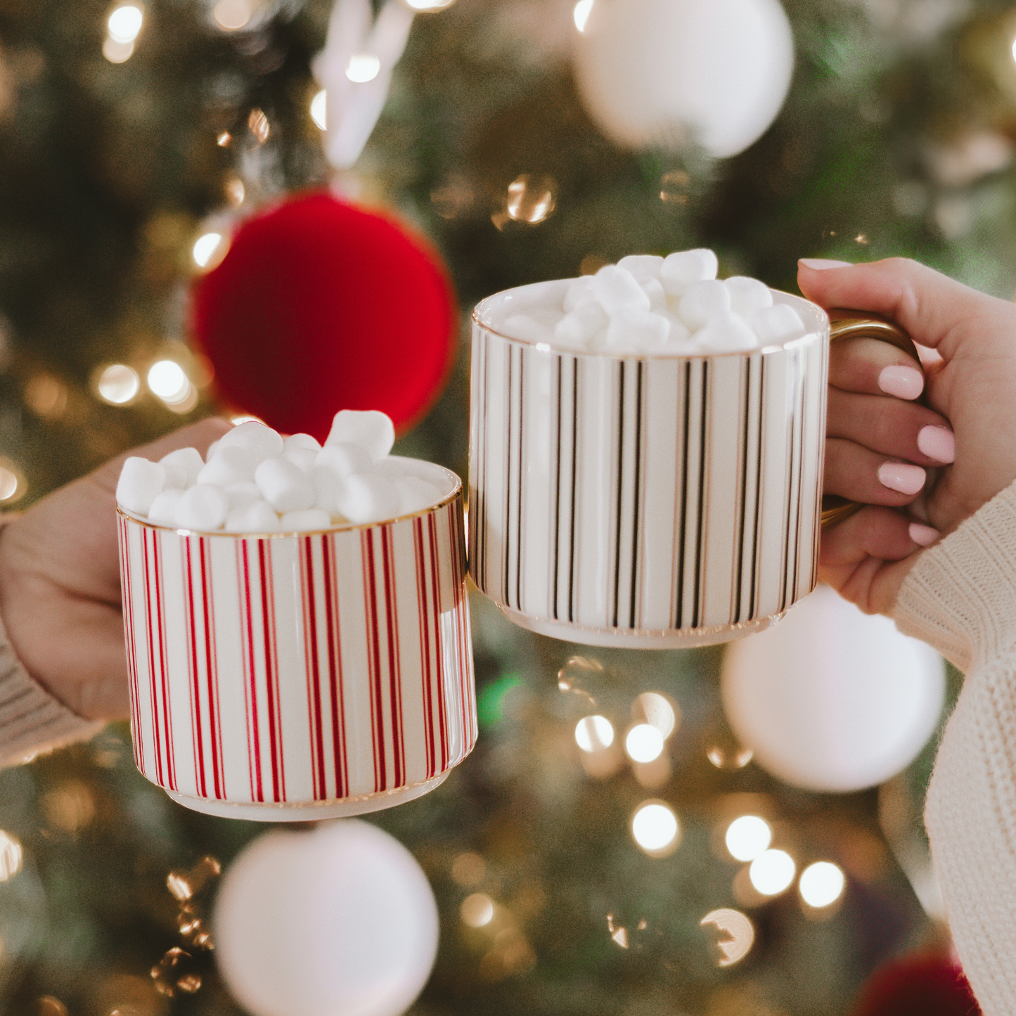 Two mugs with striped patterns filled with marshmallows, held in front of a decorated Christmas tree.