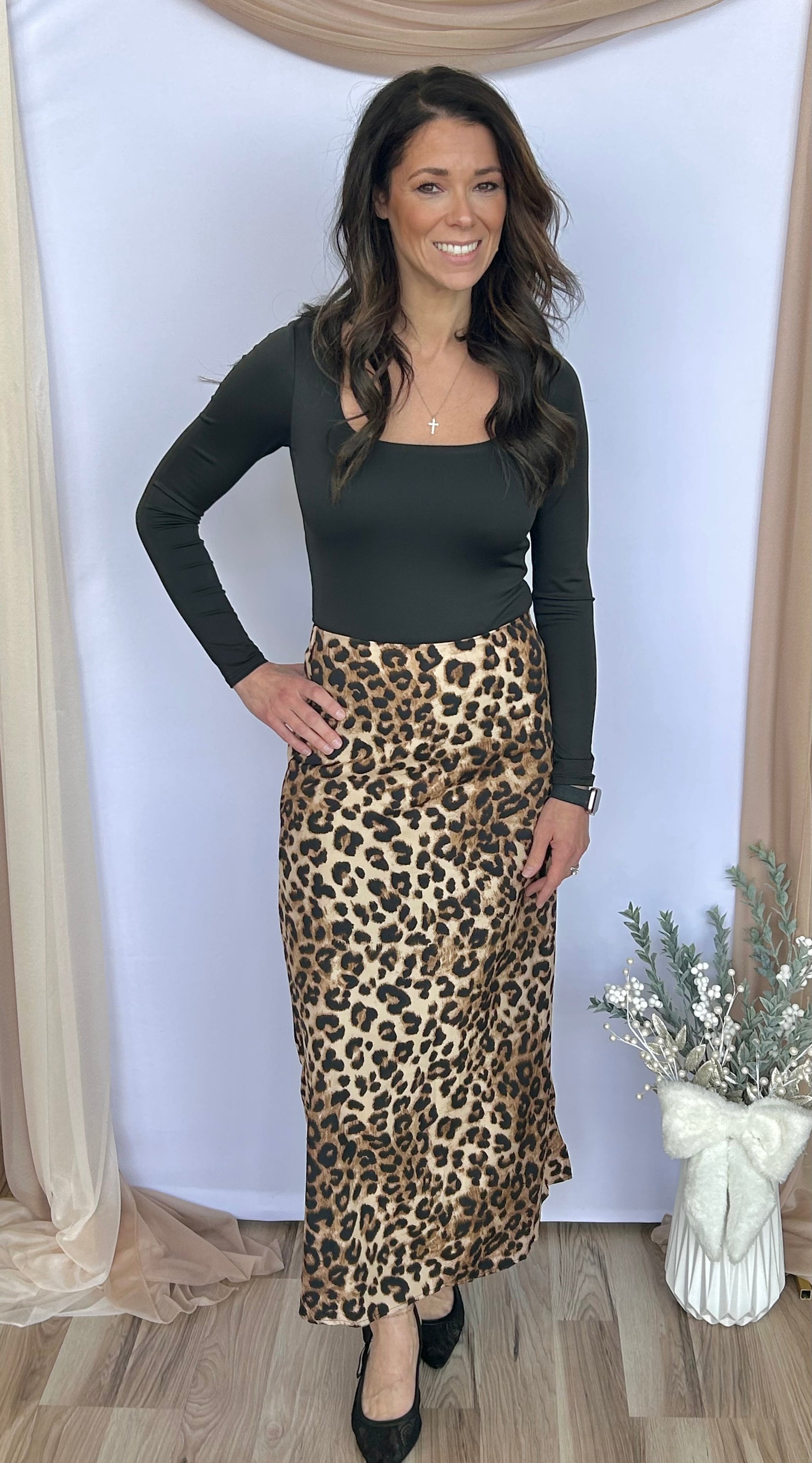 Woman wearing a black top and leopard print skirt standing against a white wall with decorative elements.