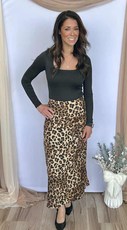 Woman wearing a black top and leopard print skirt standing against a white wall with decorative elements.