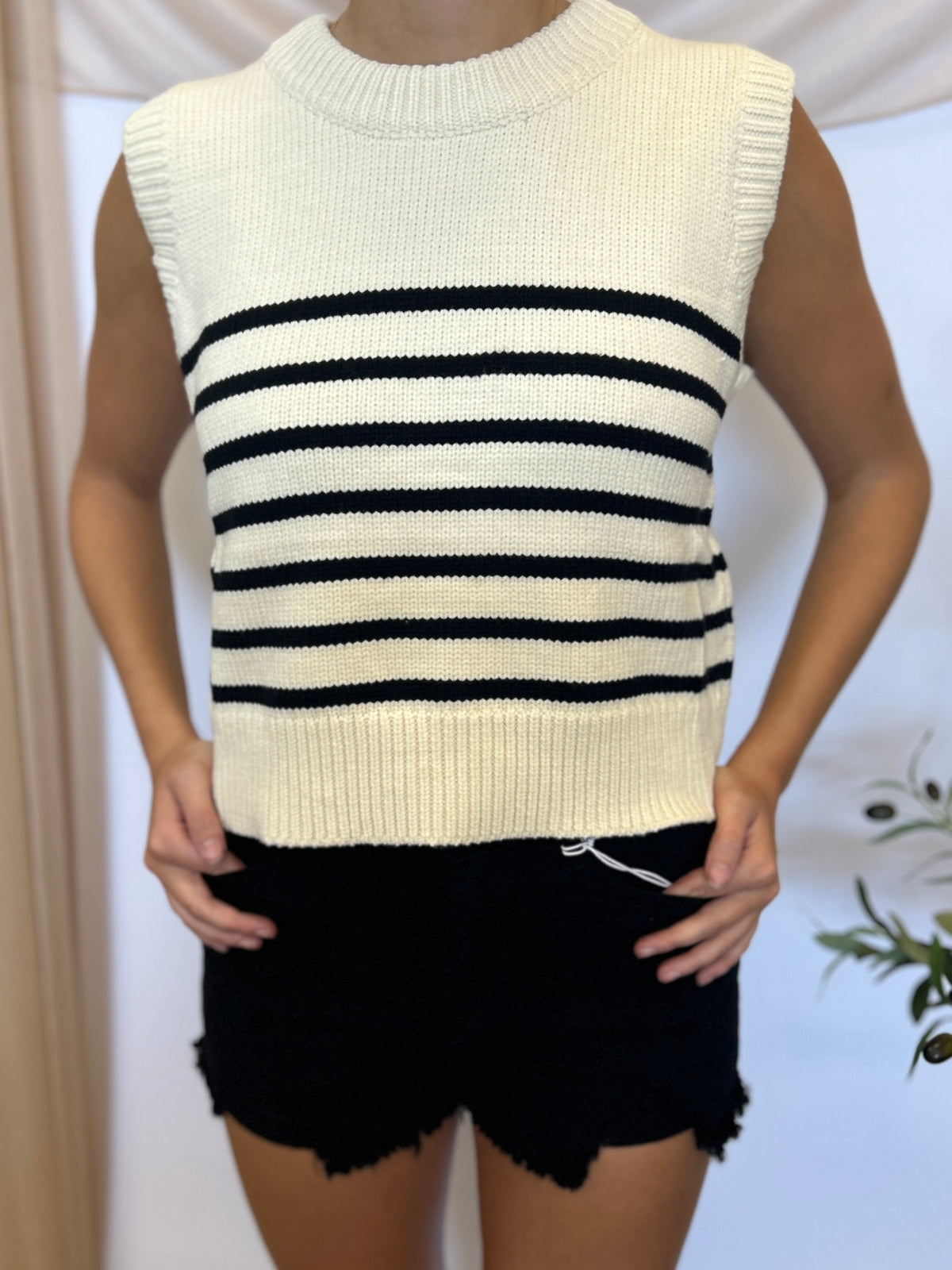 A sleeveless ribbed knit sweater vest with horizontal black stripes across an off-white base, worn by a model with black shorts.