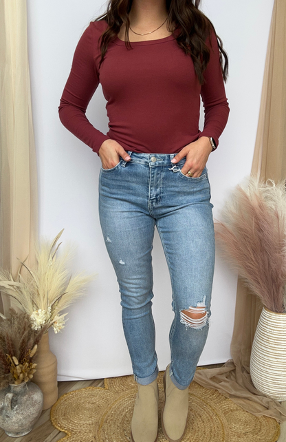 Person wearing a burgundy long-sleeve top and blue jeans standing against a white wall with decorative plants.