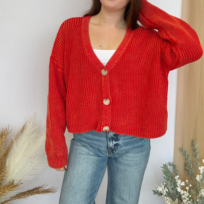 Person wearing a red cardigan over a white top and blue jeans, standing against a neutral background with decorative elements.