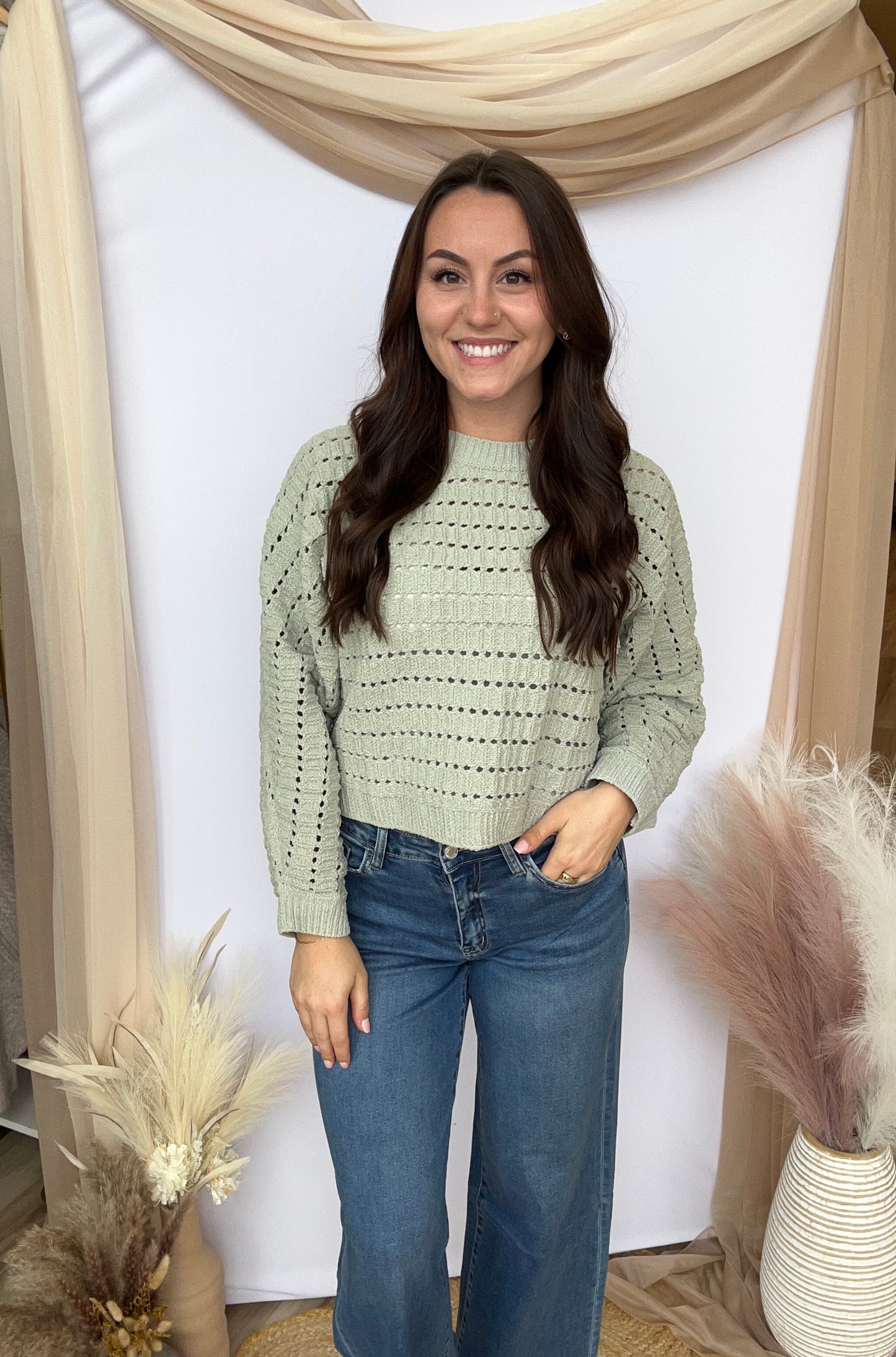 Woman wearing a light green sweater and blue jeans standing against a white wall with decorative elements.