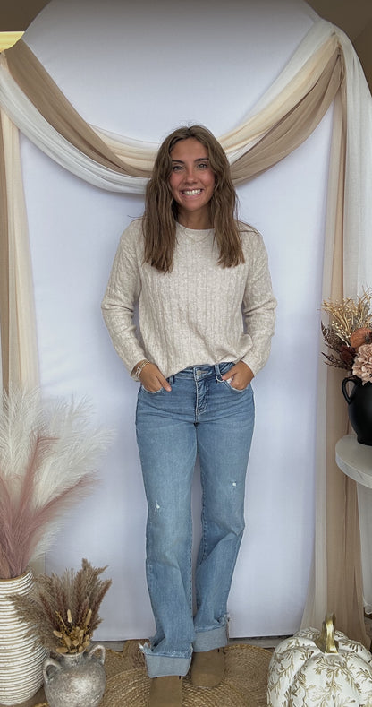 Woman wearing a beige sweater and blue jeans standing in a decorated room with plants and a mirror.
