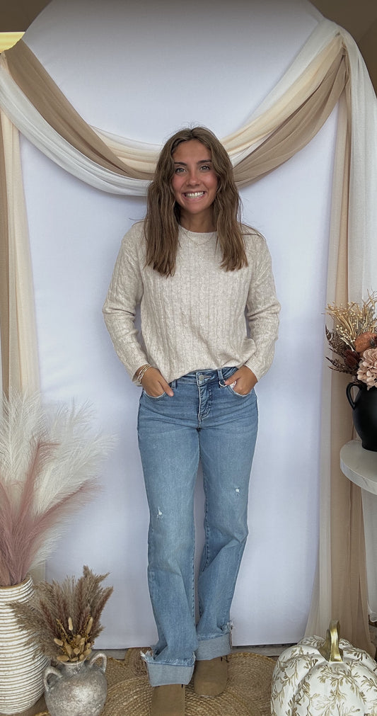 Woman wearing a beige sweater and blue jeans standing in a decorated room with plants and a mirror.