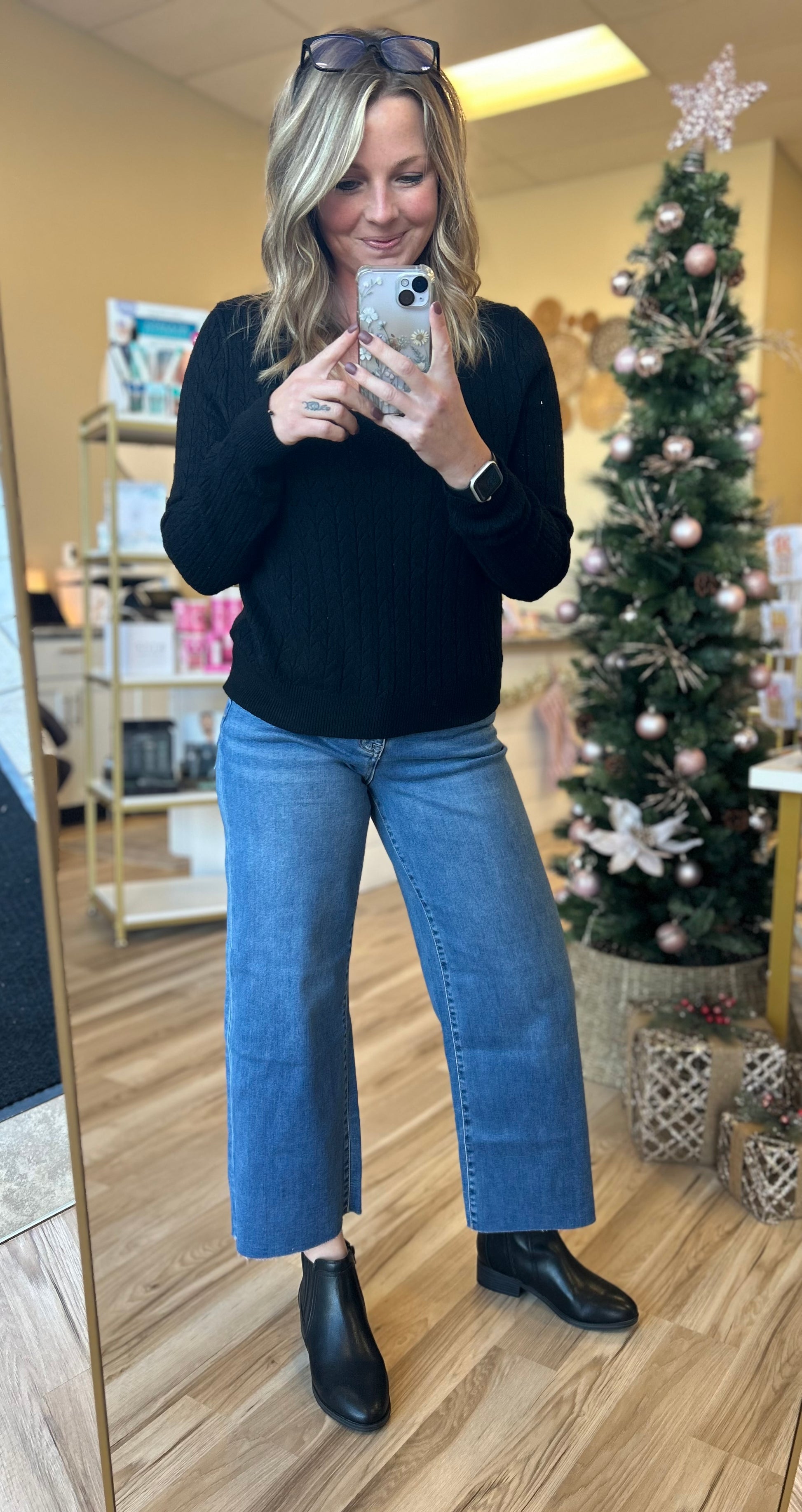 Person taking a mirror selfie wearing a black sweater and blue jeans in a store with a Christmas tree in the background.