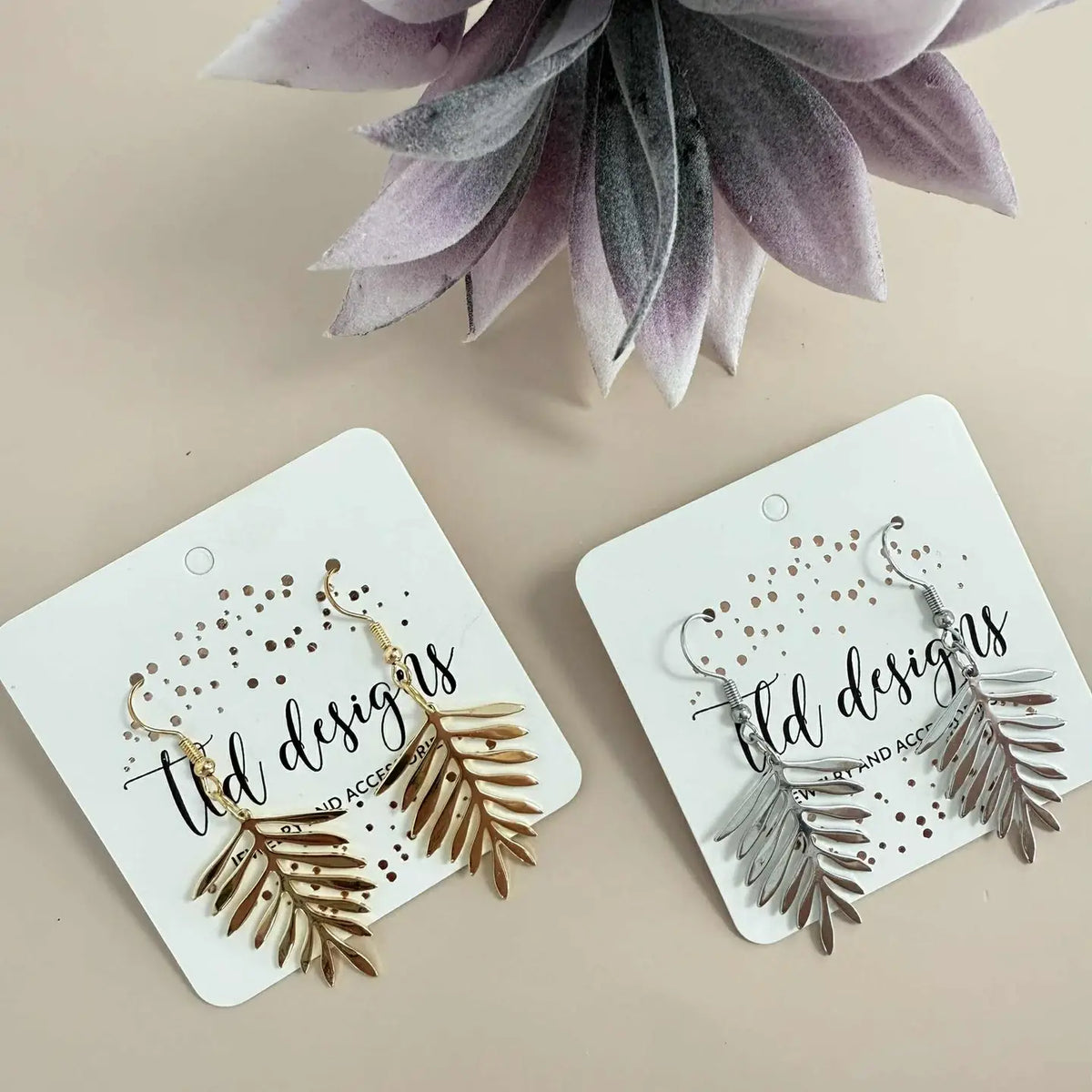 Two pairs of dangling leaf-shaped earrings displayed on white cards. One pair is gold-toned and the other is silver-toned, with the card reading 'old designs jewelry accessories.