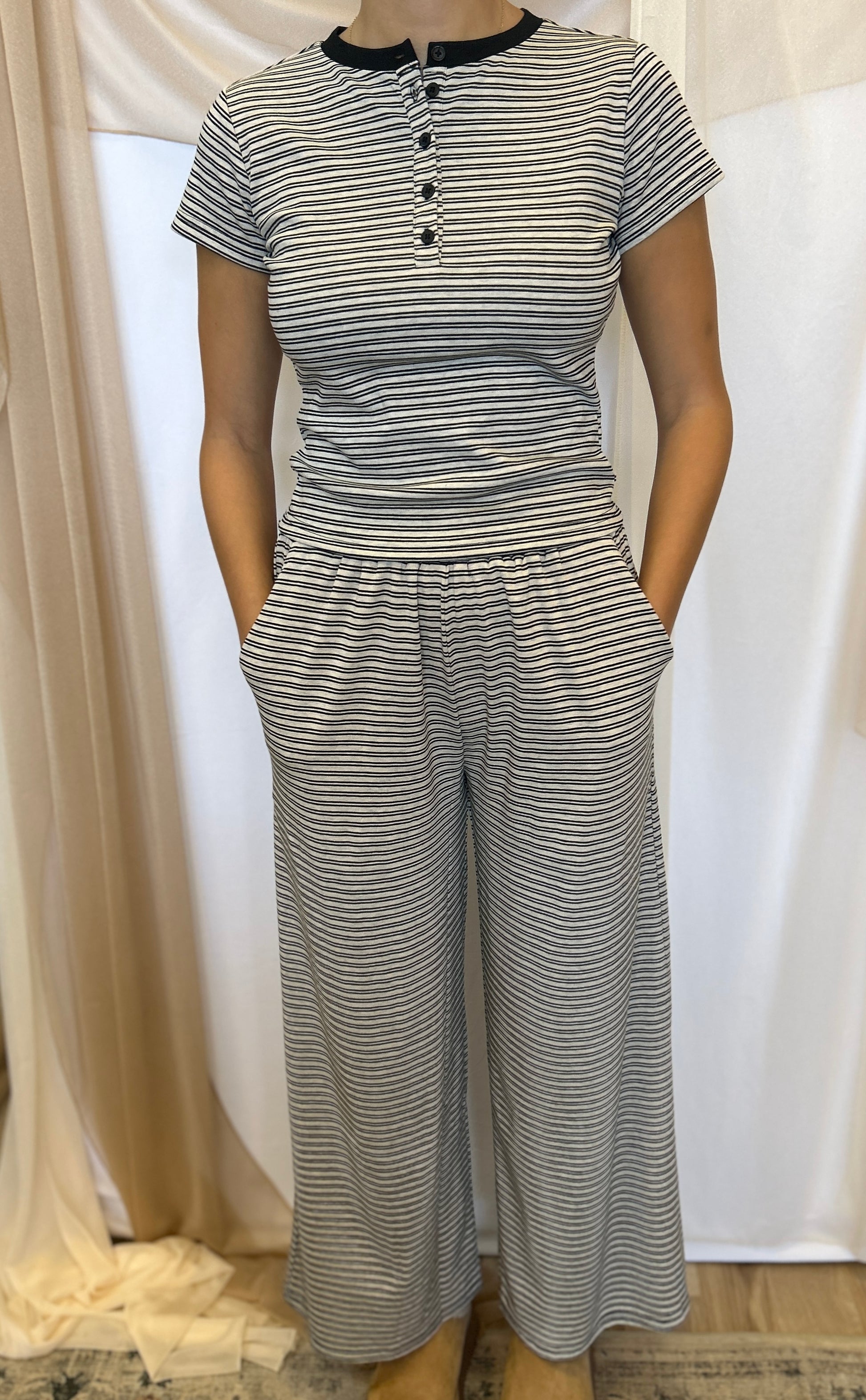 A woman wearing a matching two-piece set consisting of a short-sleeve henley top and wide-leg pants, both featuring a gray and white horizontal stripe pattern with a black collar trim on the top.