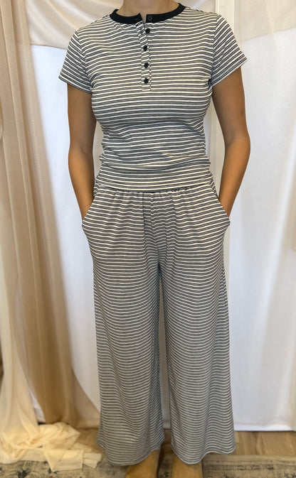 A woman wearing a matching two-piece set consisting of a short-sleeve henley top and wide-leg pants, both featuring a gray and white horizontal stripe pattern with a black collar trim on the top.