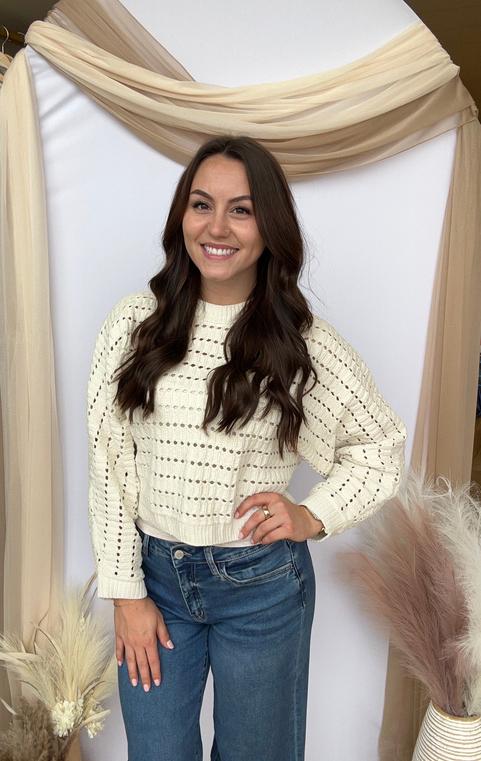 Woman wearing a white patterned sweater and blue jeans standing against a neutral backdrop.