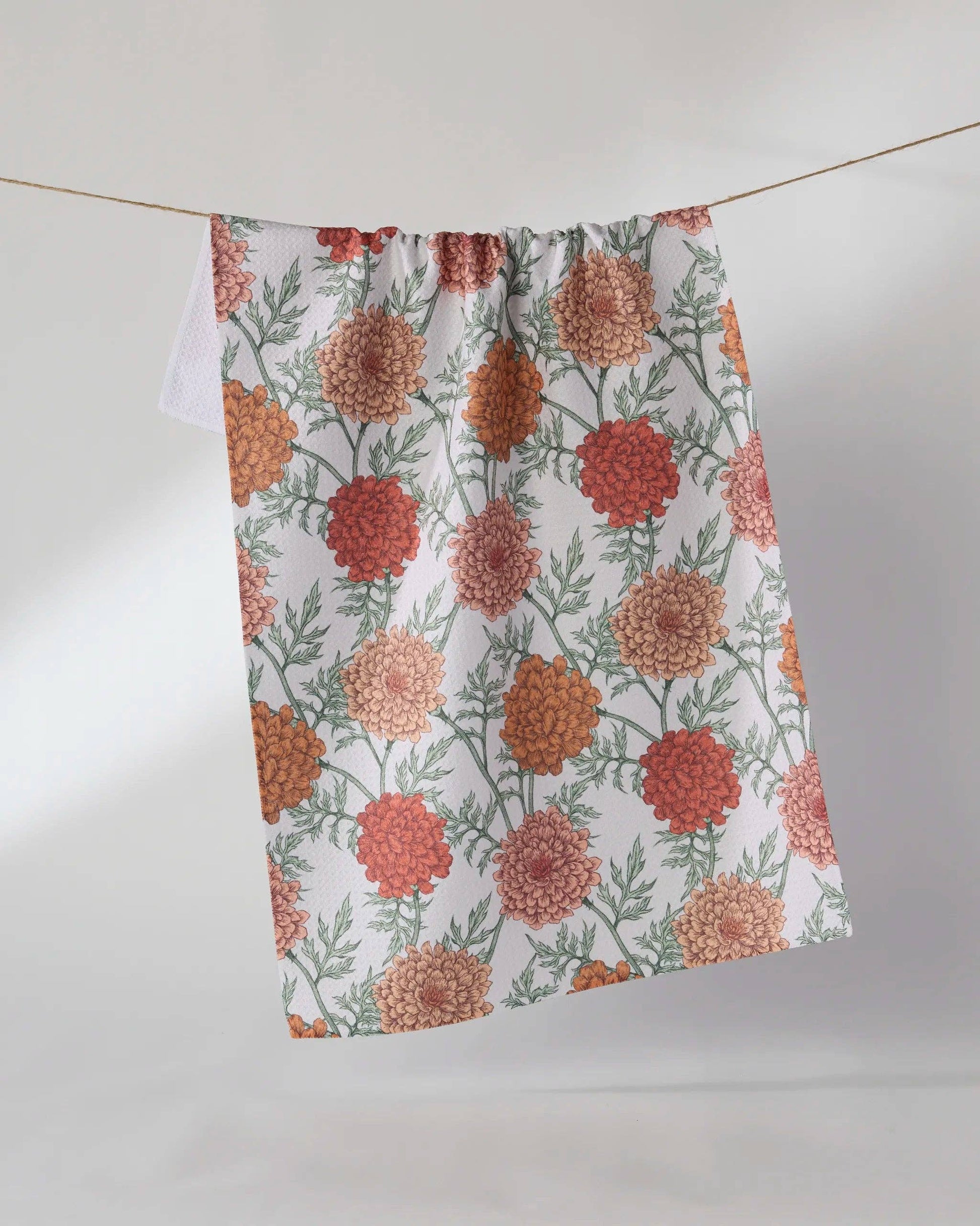 A fabric kitchen towel with a floral pattern in orange, red, and green on a white background, hanging from a string against a light gray wall.