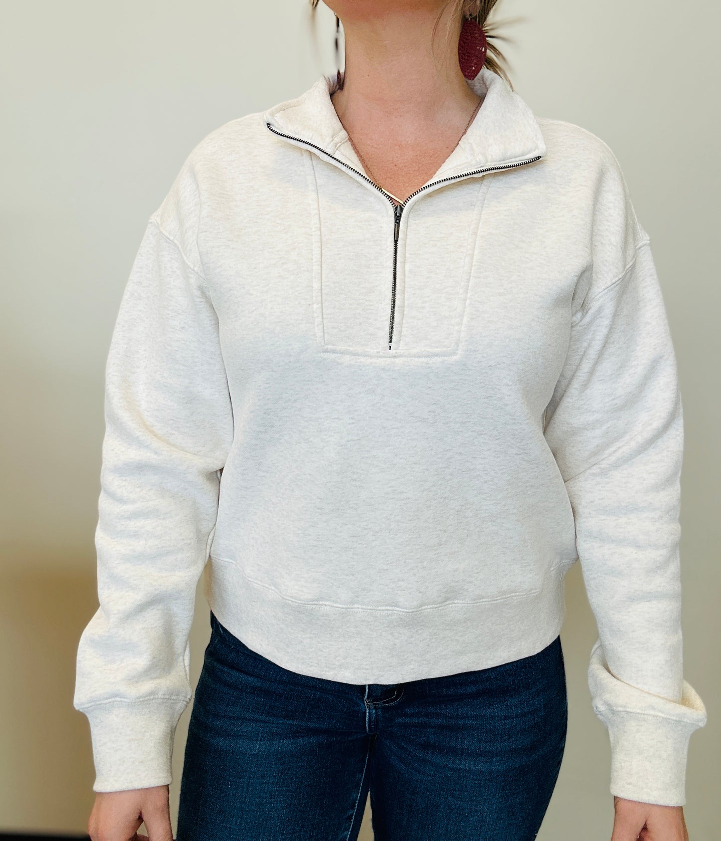 Person wearing a white pullover with a quarter zip and dark jeans against a plain background