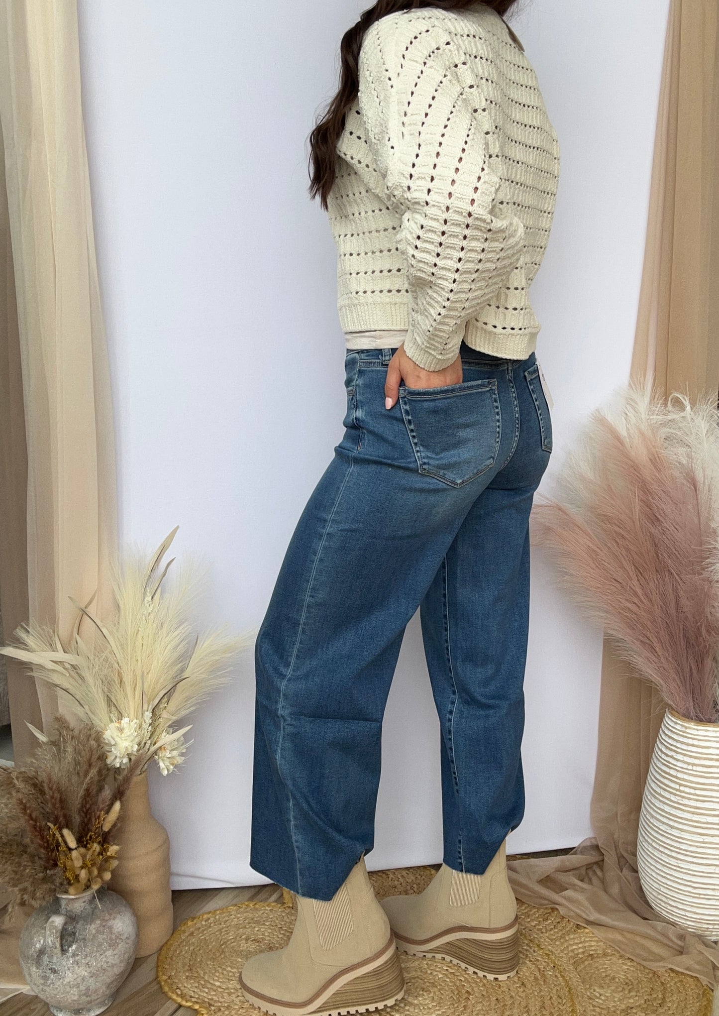 Person wearing a light-colored sweater and blue jeans standing in a room with decorative plants and a white wall.