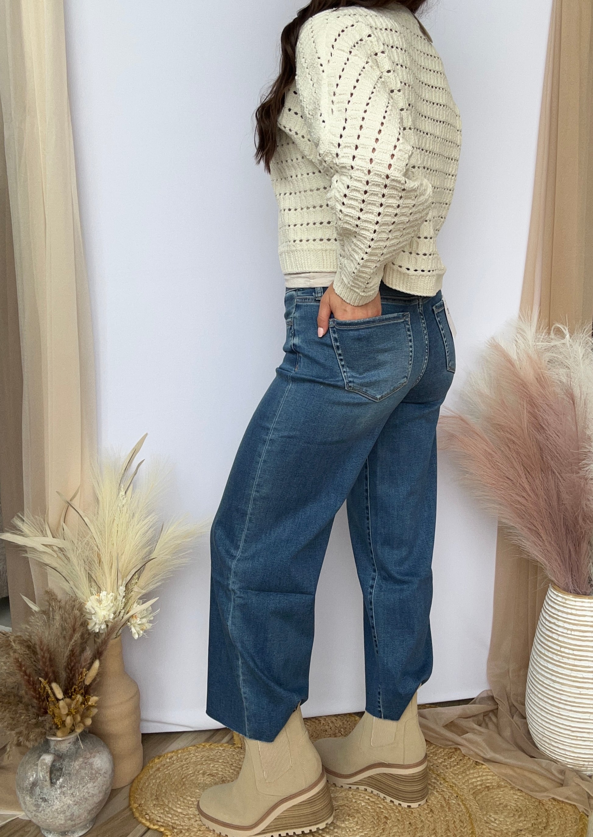Person wearing a light-colored sweater and blue jeans standing in a room with decorative plants and a white wall.