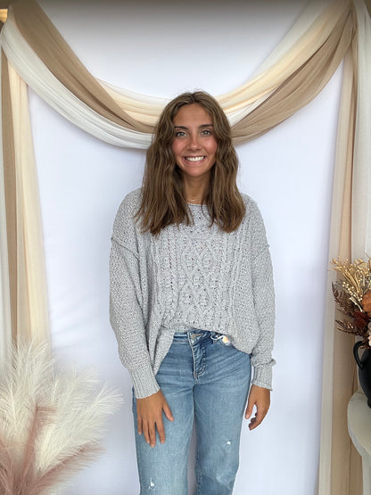 Woman wearing a gray sweater and blue jeans standing in front of a decorative wall with beige drapes.