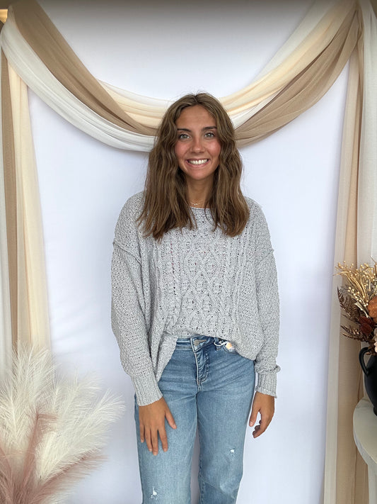 Woman wearing a gray sweater and blue jeans standing in front of a decorative wall with beige drapes.