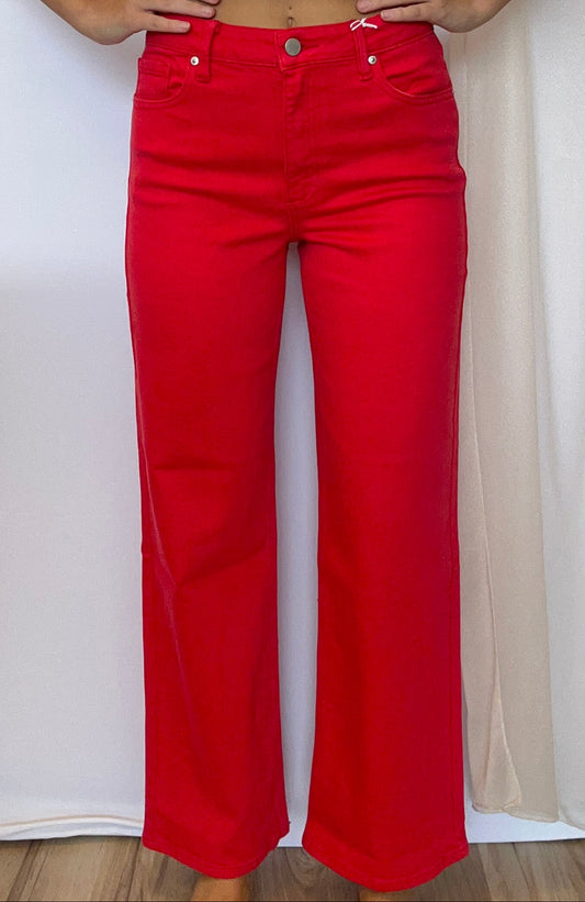 A person modeling bright red wide-leg jeans with a white ribbed short-sleeve crop top, standing against a white draped backdrop.