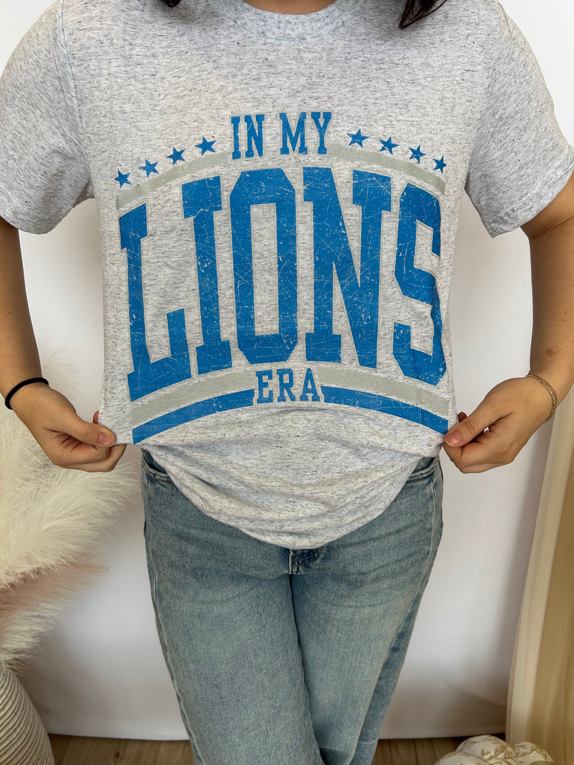 Person wearing a gray t-shirt with blue text 'IN MY LIONS ERA' and light blue jeans.