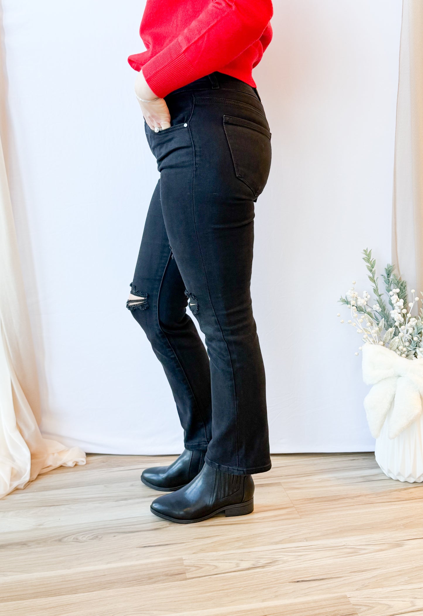 Person wearing black jeans and black boots standing on a wooden floor with a white curtain background.