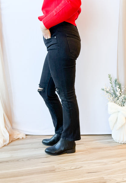 Person wearing black jeans and black boots standing on a wooden floor with a white curtain background.