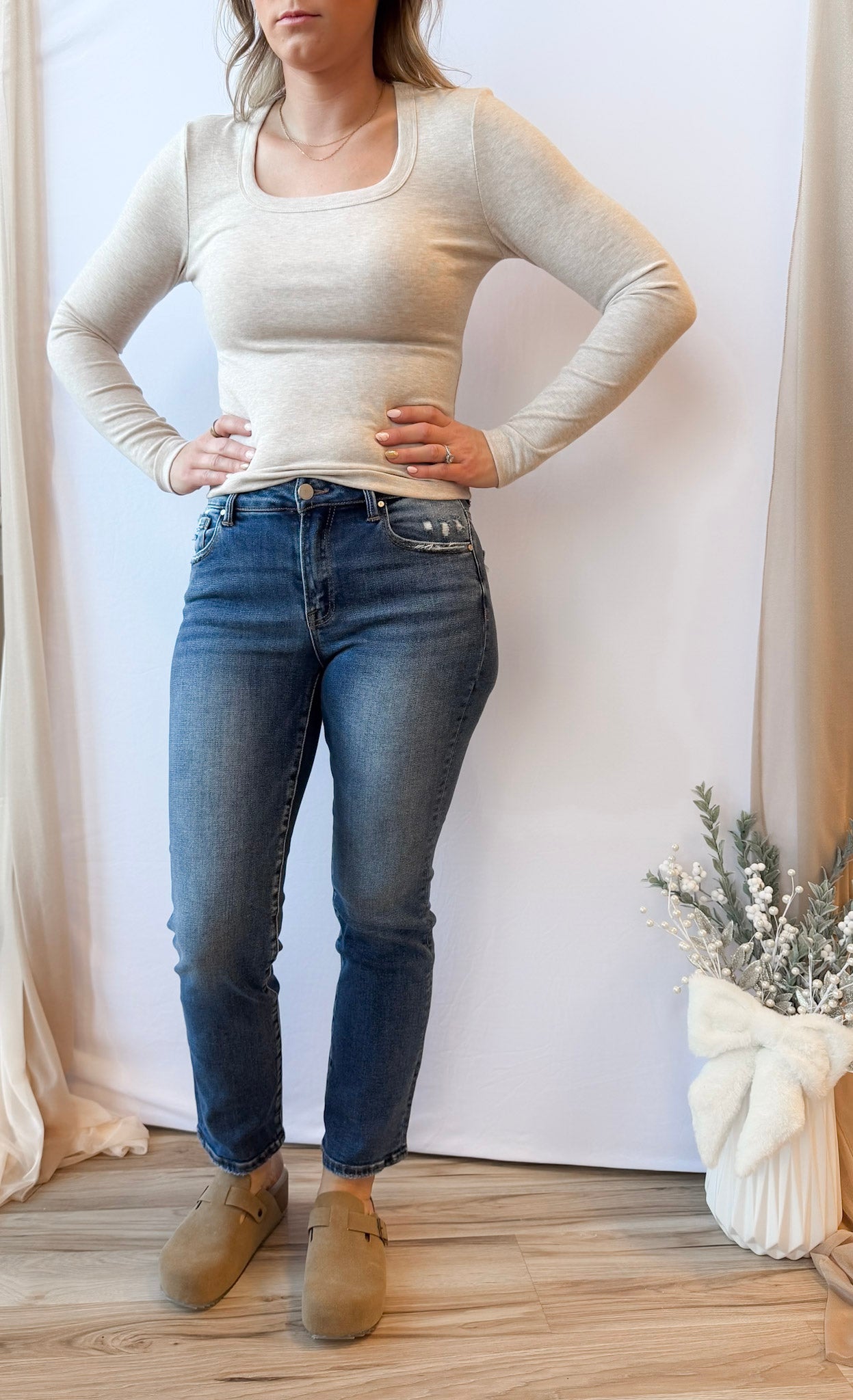 Woman wearing a beige long-sleeve top and blue jeans standing against a white wall.