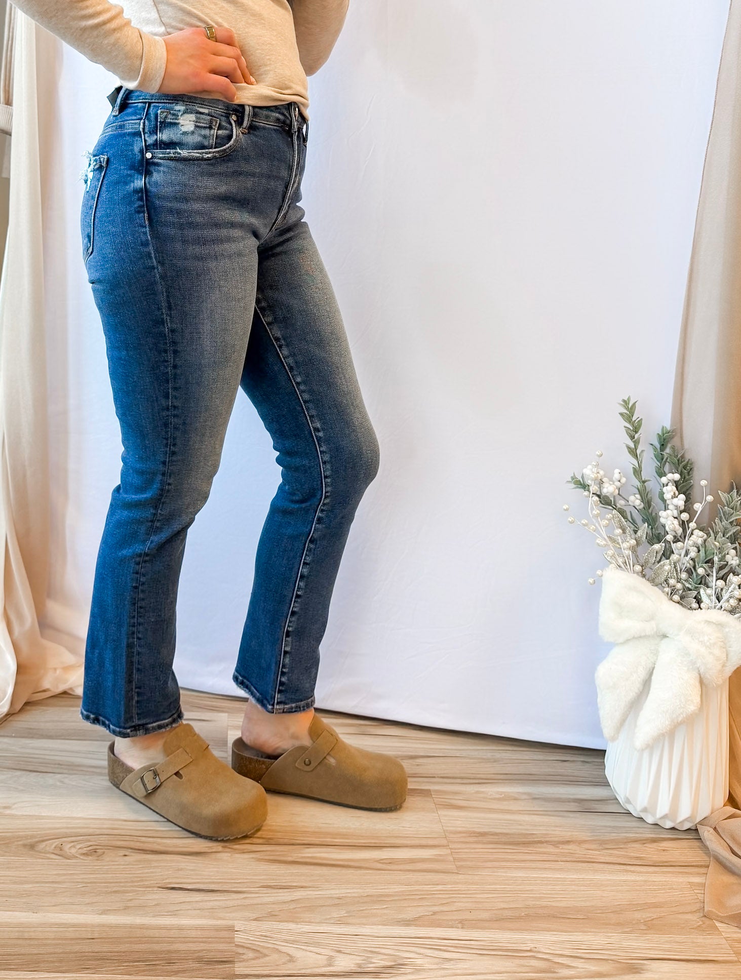 Person wearing blue jeans and brown shoes standing against a white curtain background.