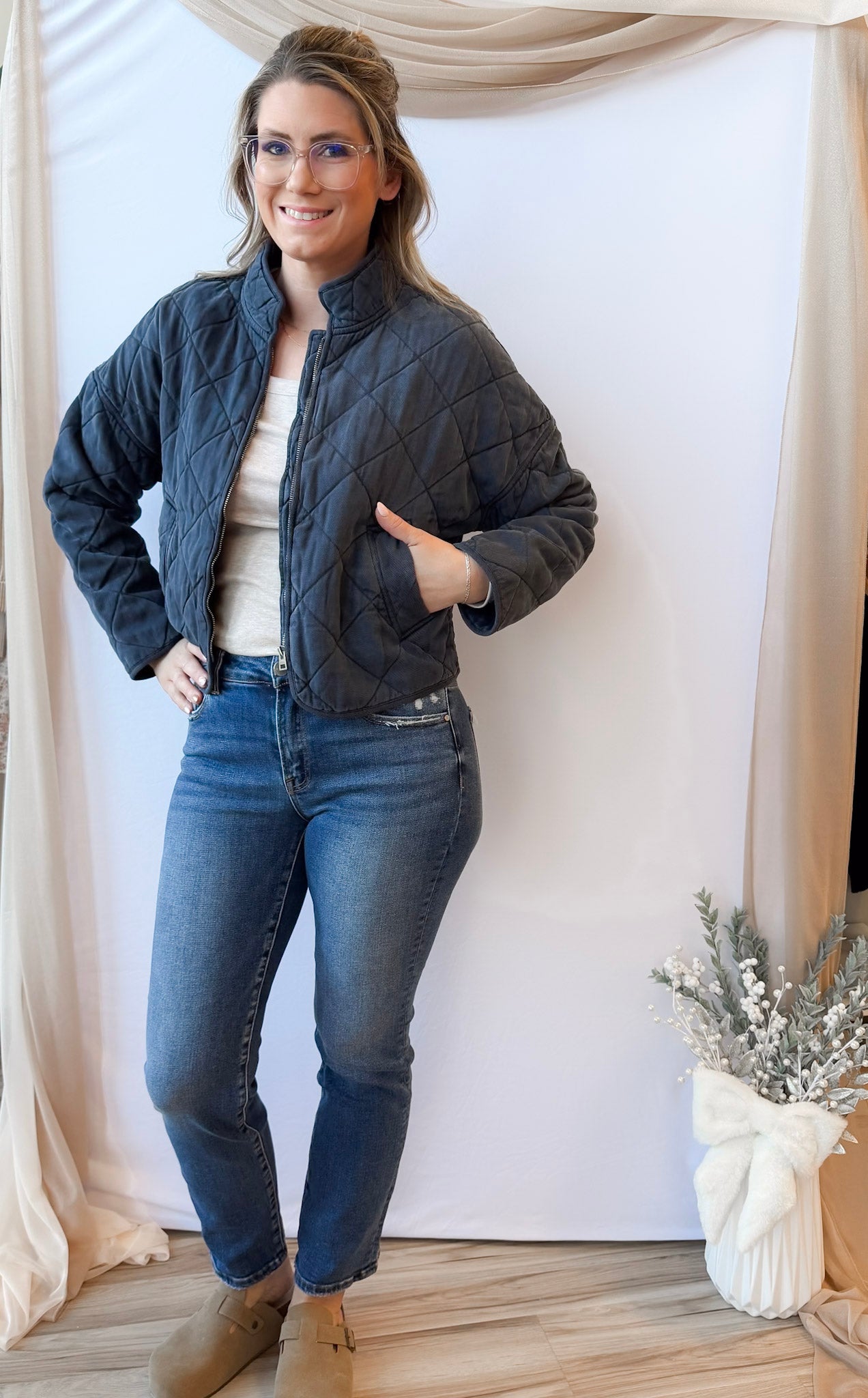 Woman wearing a dark quilted jacket and jeans standing indoors with a neutral background