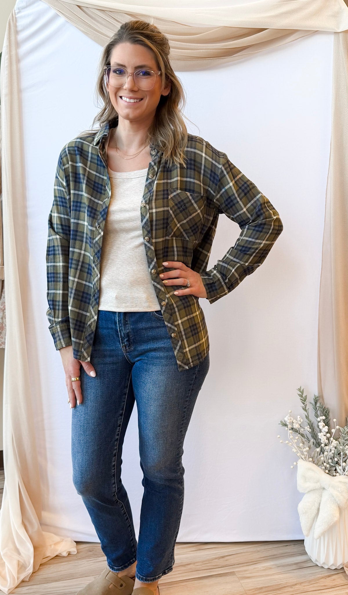 Woman wearing a plaid shirt, white top, and jeans indoors with decorative elements.