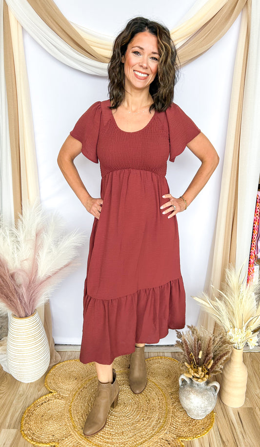 Woman wearing a burgundy dress standing in a decorated room with plants and decor.