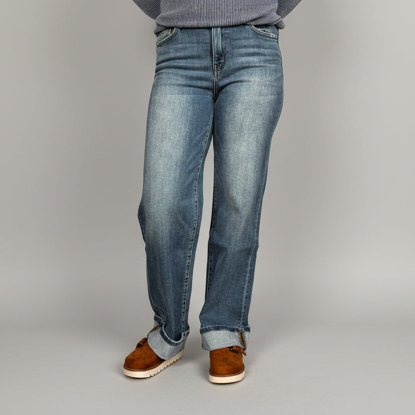 Person wearing blue jeans and brown shoes standing on a wooden floor.