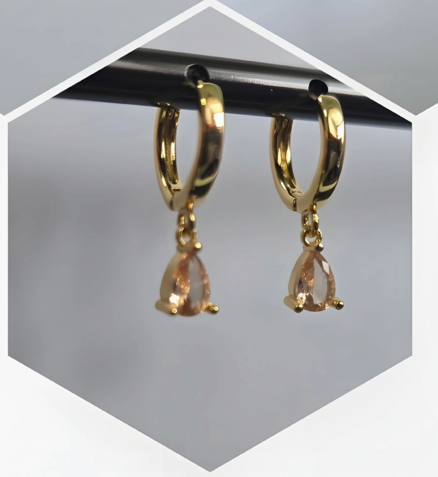 Three pairs of gold huggie hoop earrings with teardrop-shaped drop stones are displayed in a collage format. The top-left pair has a pink stone, the top-right pair has a green stone, and the bottom pair has a peach-colored stone, all shown hanging from black rods against a light gray background.