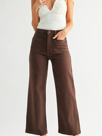 A model wearing high-waisted, wide-leg brown jeans with a white ribbed tank top, shown from the chest down against a light gray studio background.