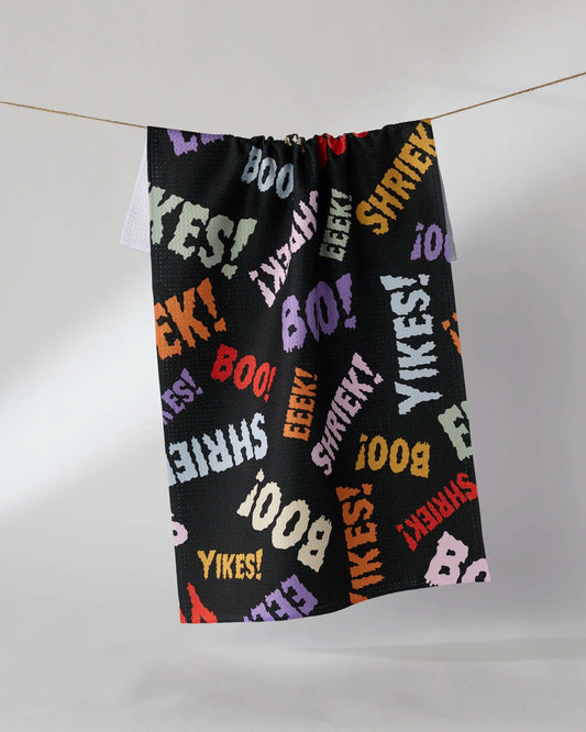 A black drawstring bag or towel-like fabric item hanging from a string against a plain light gray wall. The fabric is covered in colorful repeated text including BOO, YIKES!, EEEK!, SHRIEKS!, and LIZAH.