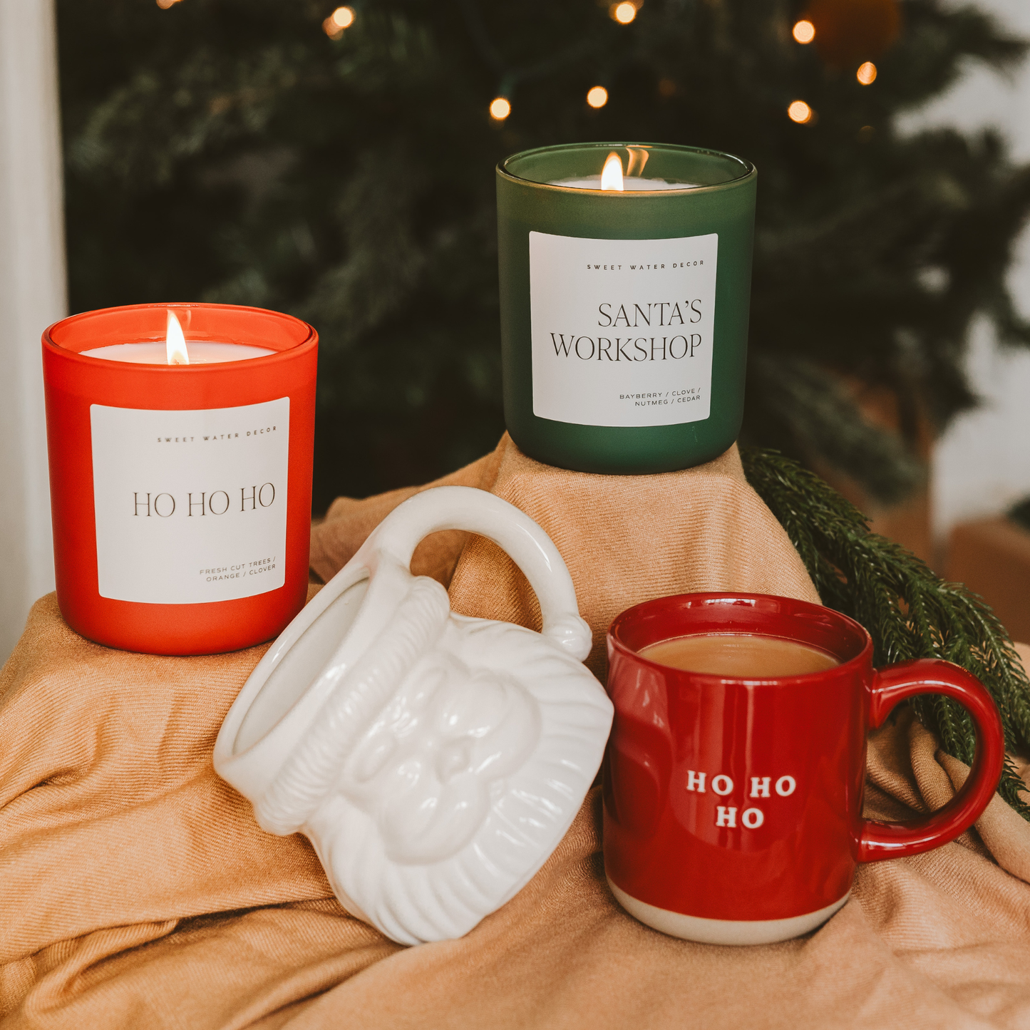 Two lit candles, a mug with 'HO HO HO', and a white container on a blanket with a Christmas tree in the background.